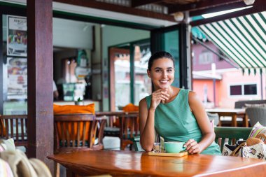 Cute elegant calm happy woman in green summer dress sits with coffee in cafe enjoying morning