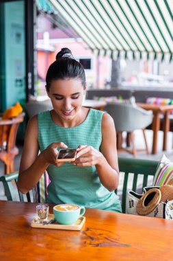 Tanned pretty woman smiling and take photo of coffee in cafe