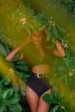 Beyaz tenli, uzun saçlı, zayıf, siyah mayo ve kemer giyen, arkasında yeşil tropikal yapraklar olan bir kadın.