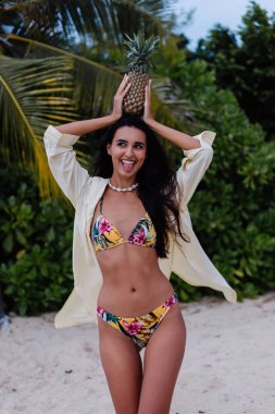 Tropikal plajda beyaz tenli, bikinili ve tişörtlü mutlu bir kadın. Egzotik ve çiçek desenli bikini.