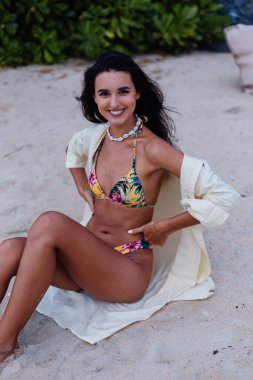 Tropikal plajda beyaz tenli, bikinili ve tişörtlü mutlu bir kadın. Egzotik ve çiçek desenli bikini.