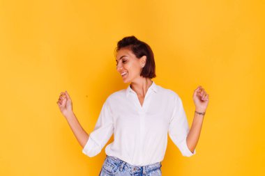 Beyaz gömlek ve kot pantolon giyen beyaz saçlı, sarı arka planda poz veren beyaz saçlı bir kadının stüdyo fotoğrafı.