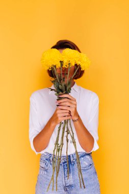 Kafkasyalı kısa saçlı, günlük kıyafetler giyen, beyaz gömlekli, kot pantolonlu, elinde bir buket sarı aster tutan mutlu bir kadın.