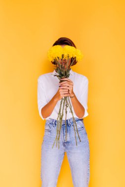 Kafkasyalı kısa saçlı, günlük kıyafetler giyen, beyaz gömlekli, kot pantolonlu, elinde bir buket sarı aster tutan mutlu bir kadın.