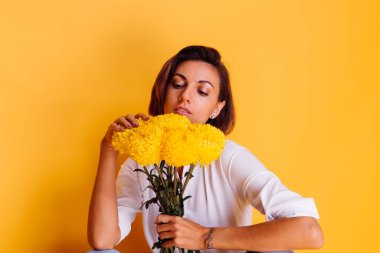 Kafkasyalı kısa saçlı, günlük kıyafetler giyen, beyaz gömlekli, kot pantolonlu, elinde bir buket sarı aster tutan mutlu bir kadın.