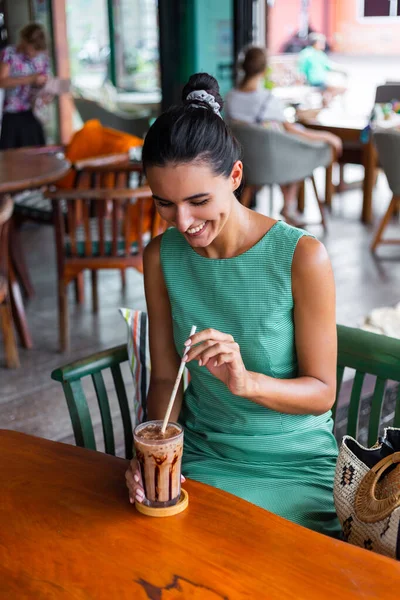 Cute elegant calm happy woman in green summer dress sits with coffee in cafe enjoying morning