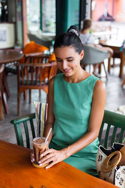 Cute elegant calm happy woman in green summer dress sits with coffee in cafe enjoying morning