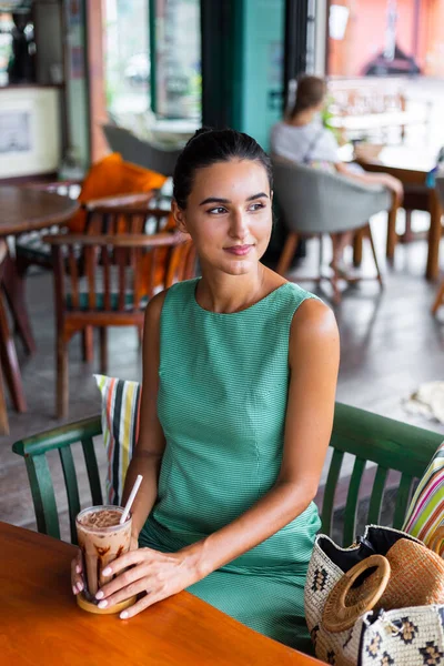 Cute elegant calm happy woman in green summer dress sits with coffee in cafe enjoying morning