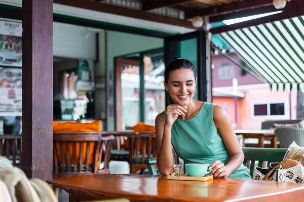 Cute elegant calm happy woman in green summer dress sits with coffee in cafe enjoying morning