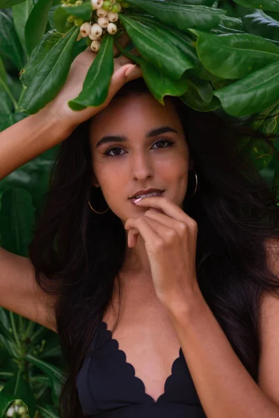 Tanned caucasian long hair fit slim woman wearing black swimsuit and belt, green tropical bushes leaves on background behind her