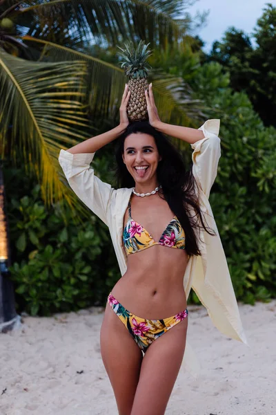 Tropikal plajda beyaz tenli, bikinili ve tişörtlü mutlu bir kadın. Egzotik ve çiçek desenli bikini.