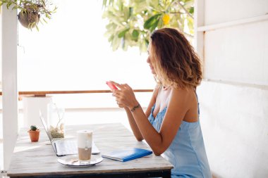 Kısa kıvırcık saçlı, romantik mavi elbiseli beyaz bir kadın açık hava kafede çalışıyor.