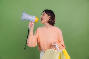 Yeşil zeytin rengi arka planda izole edilmiş şeftali rengi süveterli genç bir kadın elinde alışveriş torbalarıyla bağırıyor, indirimli satış promosyonları duyuruyor.