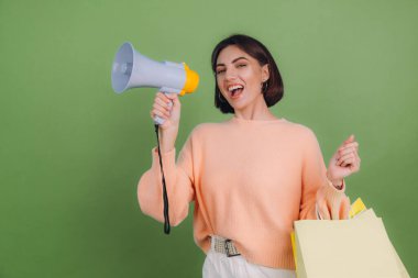 Yeşil zeytin rengi arka planda izole edilmiş şeftali rengi süveterli genç bir kadın elinde alışveriş torbalarıyla bağırıyor, indirimli satış promosyonları duyuruyor.