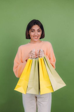 Günlük şeftali rengi süveter giyen genç bir kadın yeşil zeytin rengi arka planda izole edilmiş. Elinde alışveriş torbaları, şok edici duygular, indirimli satış promosyonu konsepti.