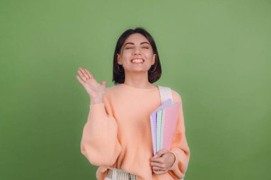 Yeşil zeytin rengi arka planda izole edilmiş gündelik şeftali rengi süveter ve sırt çantalı genç bir kadın heyecan verici duygular, açık bir eğitim konsepti.