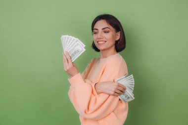 Günlük şeftali rengi süveter giyen genç bir kadın yeşil zeytin arka planda izole edilmiş. Şanslıymış ki 100 dolarlık banknotların hayranı.