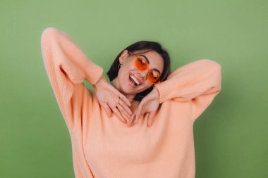 Yeşil zeytin zeminine izole edilmiş şeftali rengi süveter ve turuncu gözlüklü şık bir kadın fotokopi alanının etrafında gülümseyen mutlu bir gülüş.