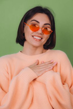 Günlük şeftali rengi süveter ve turuncu gözlüklü genç bir kadın yeşil zeytin arka planda izole edilmiş. Göğsünde el ele tutuşmuş, kalp fotokopi alanı var.