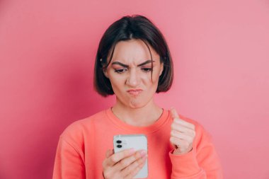 Genç bir kadın, pembe arka planda cep telefonuyla SMS okurken üzgün ve kızgın hissediyor.