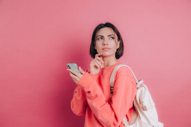 Genç, düşünceli, güzel bir kadın pembe arka planda akıllı telefonuyla düşünüyor..