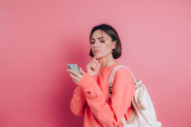 Genç, düşünceli, güzel bir kadın pembe arka planda akıllı telefonuyla düşünüyor..