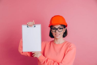 Gülümseyen kadın inşaatçı pembe arka planda beyaz tabela tutuyor. Miğfer inşa ediliyor.