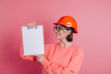Gülümseyen kadın inşaatçı pembe arka planda beyaz tabela tutuyor. Miğfer inşa ediliyor.