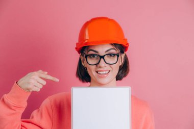 Gülümseyen kadın inşaatçı pembe arka planda beyaz tabela tutuyor. Bina kaskı. İşaret parmağı.