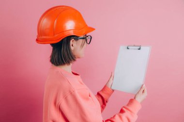 Gülümseyen kadın inşaatçı pembe arka planda beyaz tabela tutuyor. Miğfer inşa ediliyor.