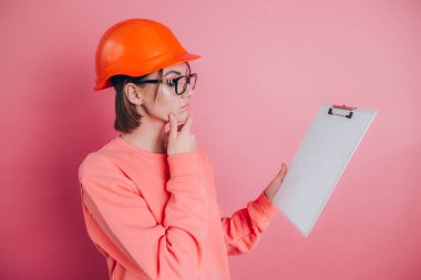 Oldukça düşünceli bir kadın müteahhit pembe arka planda beyaz tabela tutuyor. Miğfer inşa ediliyor. 