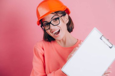 Gülümseyen kadın inşaatçı pembe arka planda beyaz tabela tutuyor. Miğfer inşa ediliyor.
