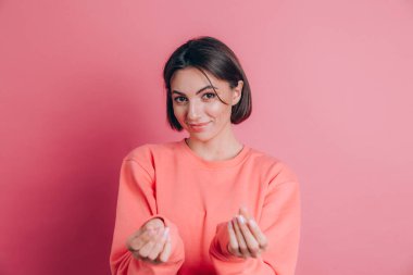 Gülümseyen genç esmer kadın, 20 'li yaşlarda, gündelik ayakta duran parmaklarını ovuşturuyor, nakit jest yapıyor, para istiyor, pembe arka plan.