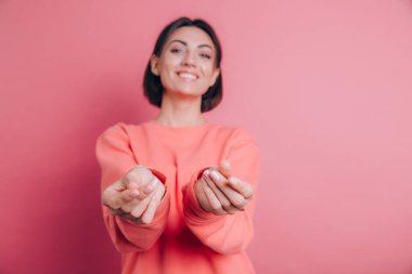 Gülümseyen genç esmer kadın, 20 'li yaşlarda, gündelik ayakta duran parmaklarını ovuşturuyor, nakit jest yapıyor, para istiyor, pembe arka plan.