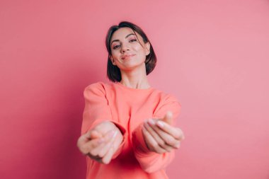 Gülümseyen genç esmer kadın, 20 'li yaşlarda, gündelik ayakta duran parmaklarını ovuşturuyor, nakit jest yapıyor, para istiyor, pembe arka plan.