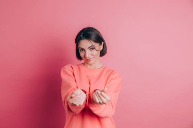 Gülümseyen genç esmer kadın, 20 'li yaşlarda, gündelik ayakta duran parmaklarını ovuşturuyor, nakit jest yapıyor, para istiyor, pembe arka plan.