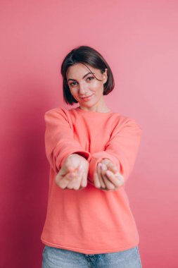 Gülümseyen genç esmer kadın, 20 'li yaşlarda, gündelik ayakta duran parmaklarını ovuşturuyor, nakit jest yapıyor, para istiyor, pembe arka plan.