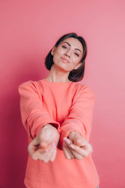 Gülümseyen genç esmer kadın, 20 'li yaşlarda, gündelik ayakta duran parmaklarını ovuşturuyor, nakit jest yapıyor, para istiyor, pembe arka plan.