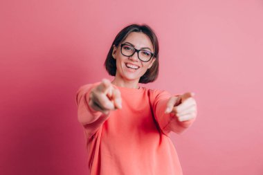 Arka planda süveter giyen kadın seni ve kamerayı parmaklarıyla gösteriyor, gülümsüyor pozitif ve neşeli.