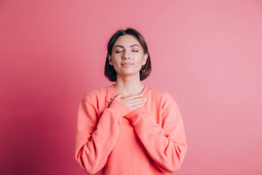 Arka planda sıradan bir süveter giyen, elleri göğsünde, gözleri kapalı ve yüzünde minnet dolu bir jest olan bir kadın.