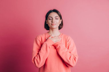 Arka planda sıradan bir süveter giyen, elleri göğsünde, gözleri kapalı ve yüzünde minnet dolu bir jest olan bir kadın.