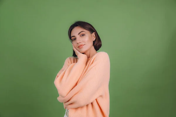 Young woman in casual peach sweater isolated on green olive color background love concept, holding hands crossed hugging herself