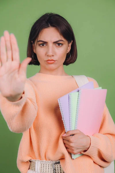 Young woman in casual peach sweater isolated on green olive color background with notebooks showing stop gesture, education high school concept