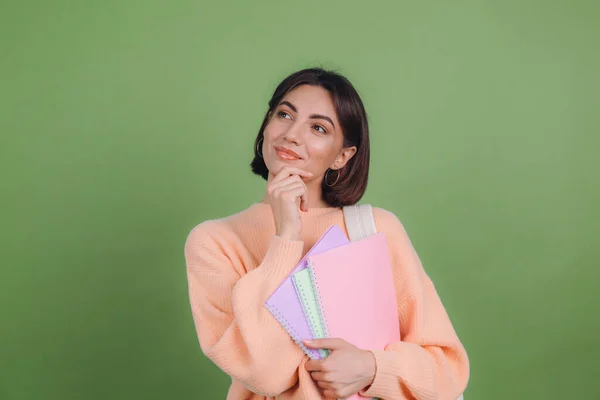 Young woman in casual peach sweater isolated on green olive color background with notebooks thoughtful positive touch chin with hand, education high school concept