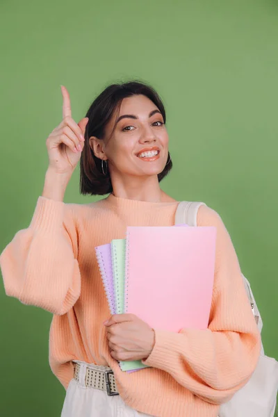 Young woman in casual peach sweater isolated on green olive color background with notebooks excited pointing finger up, idea came up, education high school concept