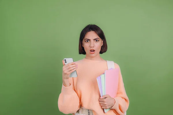 Young woman in casual peach sweater and backpack isolated on green olive color background with notebooks shocked with mobile phone education high school concept