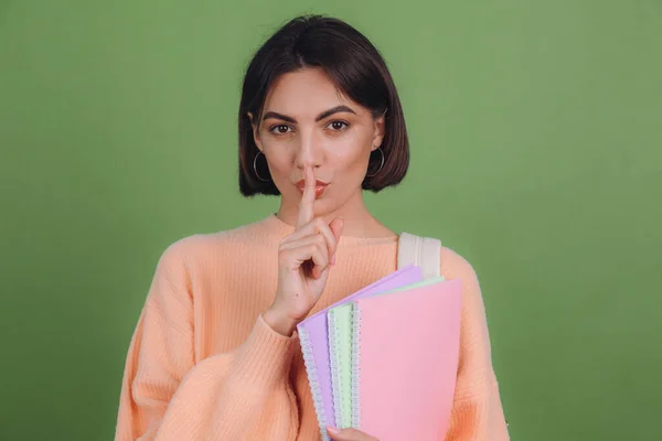 Young woman in casual peach sweater and backpack isolated on green olive color background say be quiet with finger on lips shhh secret gesture education concept