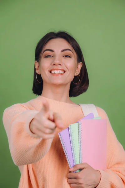 Young woman in casual peach sweater and backpack isolated on green olive color background smiling happy pointing finger to camera education concept