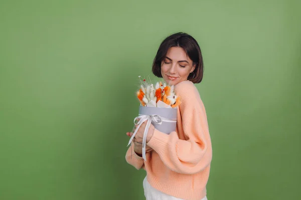 Young woman in casual peach sweater  isolated on green olive background  hold  orange white flower box composition of cotton flowers, gypsophila, wheat and lagurus for a gift happy amazed surprised face