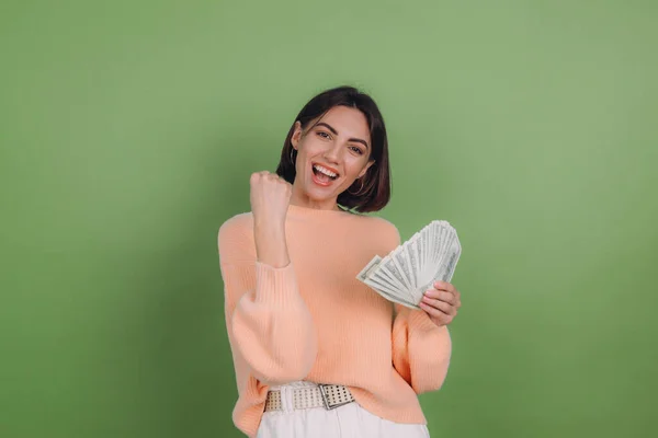 Young woman in casual peach sweater  isolated on green olive background  lucky holding fan of 100 dollar bills clenching fist winner gesture copy space